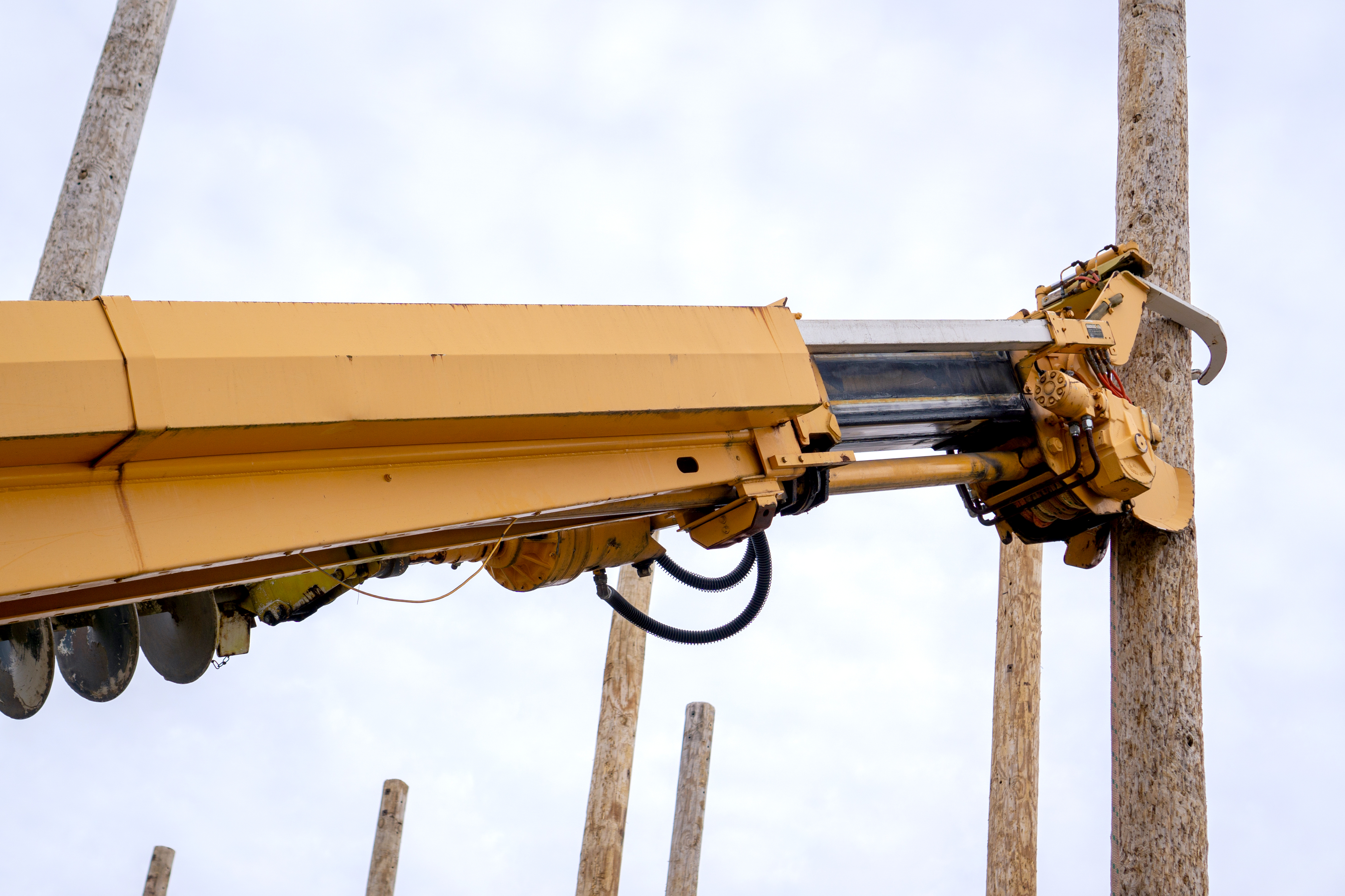 Yellow digger truck arm with claws clamped around wooden utility power line pole against cloudy sky with poles in background